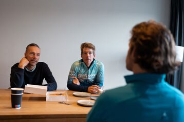 Drie mannen zitten aan een tafel met koffie en gebak.