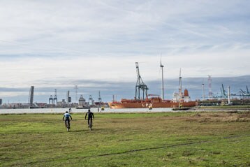 Twee fietsers bij een industrieel havengebied met kranen en een vrachtschip op de achtergrond.