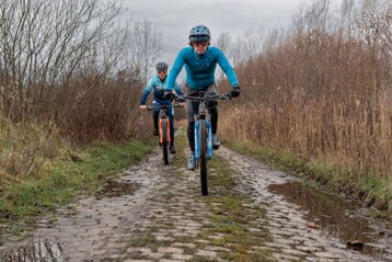 Twee fietsers rijden op een modderig kasseienpad door de natuur.