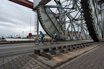 Fietsers rijden onder een grote industriële constructie bij een waterkant.