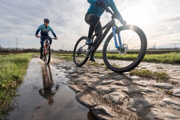 Twee fietsers rijden op een kasseienpad met een plas, één fietser op de voorgrond.