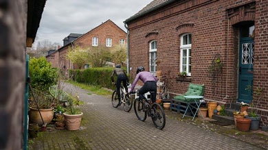 Twee fietsers rijden door een steegje met klinkers langs planten en rode bakstenen gebouwen.