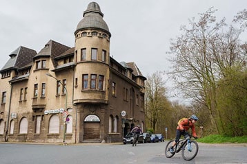 Fietsers rijden langs een oud gebouw met een torentje in een met bomen omzoomde straat.