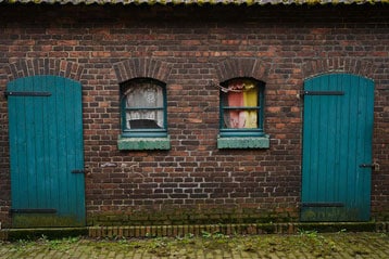Bakstenen muur met twee boogvensters en turquoise deuren.