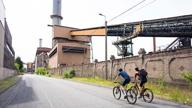 Twee fietsers rijden langs industriële gebouwen.