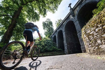Fietser rijdt bergopwaarts richting een stenen brug omgeven door bomen.