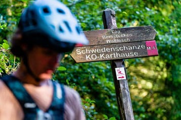 Fietser bij een houten wandelpadbord in een bos.