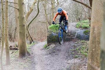 Fietser in oranje trui springt over een boomstam op een bospad.
