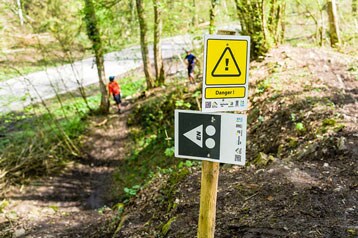Pad met waarschuwingsborden, bos op de achtergrond, twee wandelaars in de verte.