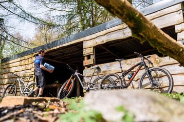 Persoon met een container bij een houten schuur en fietsen in het bos.
