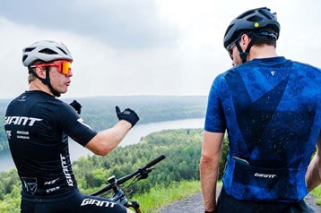Twee fietsers met helmen en jerseys kijken uit over een schilderachtig meer en bos.