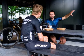 Twee fietsers in een café in gesprek met eten en fietsen in de buurt.