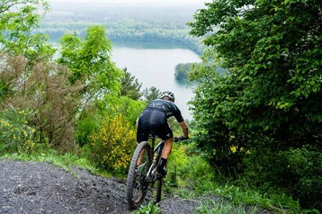 Fietser rijdt bergafwaarts op een pad met uitzicht op een meer en weelderig groen.