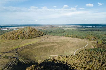 Luchtfoto van een grasrijk landschap met heuvels en bossen onder een blauwe lucht.