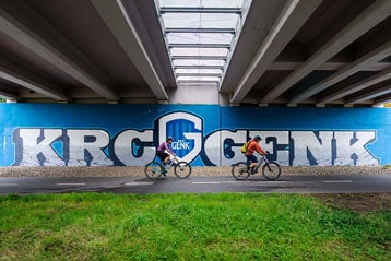 Fietsers rijden langs een KRC Genk-muurschildering onder een brug.
