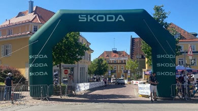 Ein grünes aufblasbares Tor mit „Skoda“-Branding überspannt eine sonnige Stadtstraße, umgeben von Bäumen, Gebäuden und Menschen, die sich für eine Outdoor-Veranstaltung versammeln.