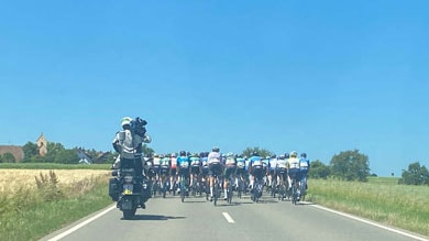Ein Peloton von Radfahrern fährt auf einer geraden Landstraße unter klarem blauem Himmel. Ein Kameramann auf einem Motorrad folgt dicht dahinter, um das Rennen aufzunehmen.