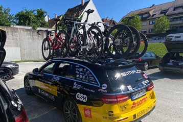 Ein schwarz-gelbes Teamfahrzeug mit mehreren Rennrädern auf dem Dachgepäckträger steht auf einem sonnigen Parkplatz. Im Hintergrund sind Gebäude mit Bäumen zu sehen.