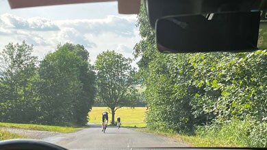 Blick aus einem Auto, das zwei Radfahrern auf einer ländlichen Straße folgt, mit üppigen grünen Bäumen und Feldern im Hintergrund unter einem teilweise bewölkten Himmel.