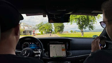 Im Begleitfahrzeug eines Radrennens überwachen Fahrer und Beifahrer das Rennen mit einem GPS-Navigationsbildschirm, während Radfahrer auf einer offenen Straße vorausfahren.