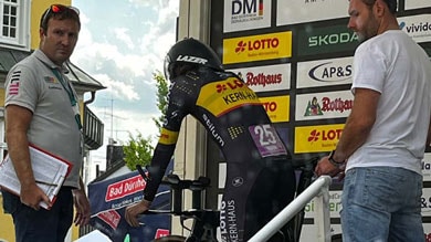 Ein Radfahrer im schwarzen und gelben Skinsuit bereitet sich auf den Start eines Zeitfahrens vor, steht auf einer Rampe mit zwei Offiziellen daneben. Sponsor-Banner sind im Hintergrund zu sehen.
