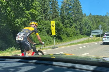 Ein Radfahrer im schwarz-gelben Trikot fährt neben einem Begleitfahrzeug an einem 'Start Waste Zone'-Schild vorbei. Die Straße ist von Bäumen gesäumt, Teamfahrzeuge sind voraus.