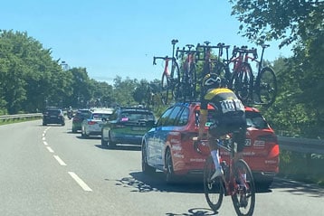 Ein Radfahrer mit der Startnummer 131 fährt hinter einem roten Teamwagen mit transportierten Fahrrädern auf einer sonnigen Straße, umgeben von Bäumen und anderen Fahrzeugen im Rennkonvoi.