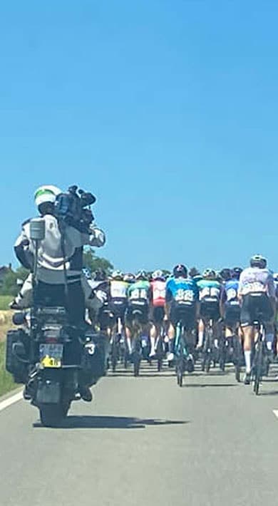 Eine Gruppe von Radfahrern fährt dicht beieinander auf einer ländlichen Straße unter klarem blauem Himmel, gefolgt von einem Kameramann auf einem Motorrad, der das Ereignis festhält.