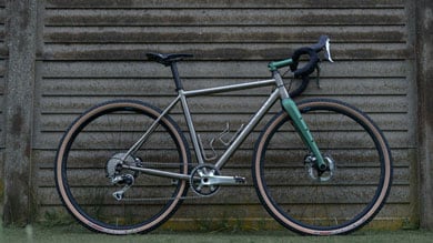 Grey road bike with green fork and drop handlebars, leaning against a ribbed concrete wall.