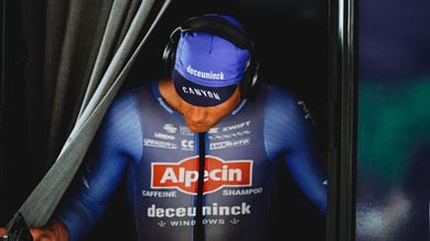 Wielrenner in een blauw shirt en een pet, draagt een koptelefoon en loopt door een gordijn.