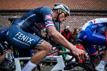 Wielrenner in een blauw shirt rijdt in een wedstrijd met toeschouwers op de achtergrond.