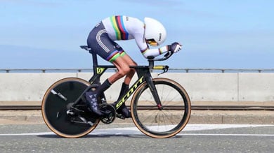 Radfahrer im regenbogen-gestreiften Trikot, gebeugt über ein schwarzes Zeitfahrrad, mit stromlinienförmigem Helm, fährt auf einer Straße gegen einen klaren blauen Himmel.