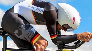 Ein Radfahrer in einem aerodynamischen schwarz-weiß-roten deutschen Trikot fährt ein Zeitfahrrad vor blauem Himmel. Die fokussierte und intensive Haltung deutet auf Geschwindigkeit hin.