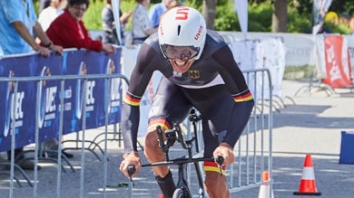 Ein Radfahrer in schwarzer und roter Ausrüstung mit Helm fährt ein Zeitfahrrad auf einer Strecke mit Absperrungen. Zuschauer beobachten ihn, während er an einem sonnigen Tag Tempo macht.