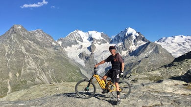 Eine Person steht mit einem Mountainbike auf einem felsigen Plateau und lächelt vor einer Kulisse aus imposanten, schneebedeckten Gipfeln und klarem blauen Himmel.