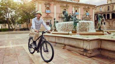 Person riding a bicycle near a fountain in a European plaza with historic buildings in the background