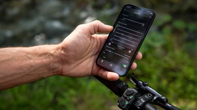 Hand holding smartphone displaying cycling app settings, near bicycle handlebars in an outdoor environment