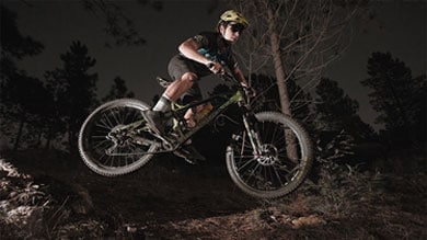 Um ciclista de montanha executa um salto no ar à noite, iluminado por luzes dramáticas. Com capacete e equipamento de proteção, ele sobrevoa um trilho de terra em meio a uma área arborizada.