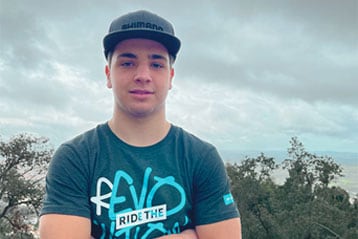 Um jovem com boné Shimano e t-shirt cruza os braços contra uma paisagem ao ar livre. Com expressão confiante, ele posa diante de um céu nublado e vegetação exuberante que se estende ao fundo.