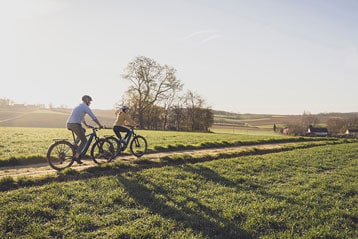 Two people riding bicycles on a rural path in a scenic landscape with fields and trees under a bright sky