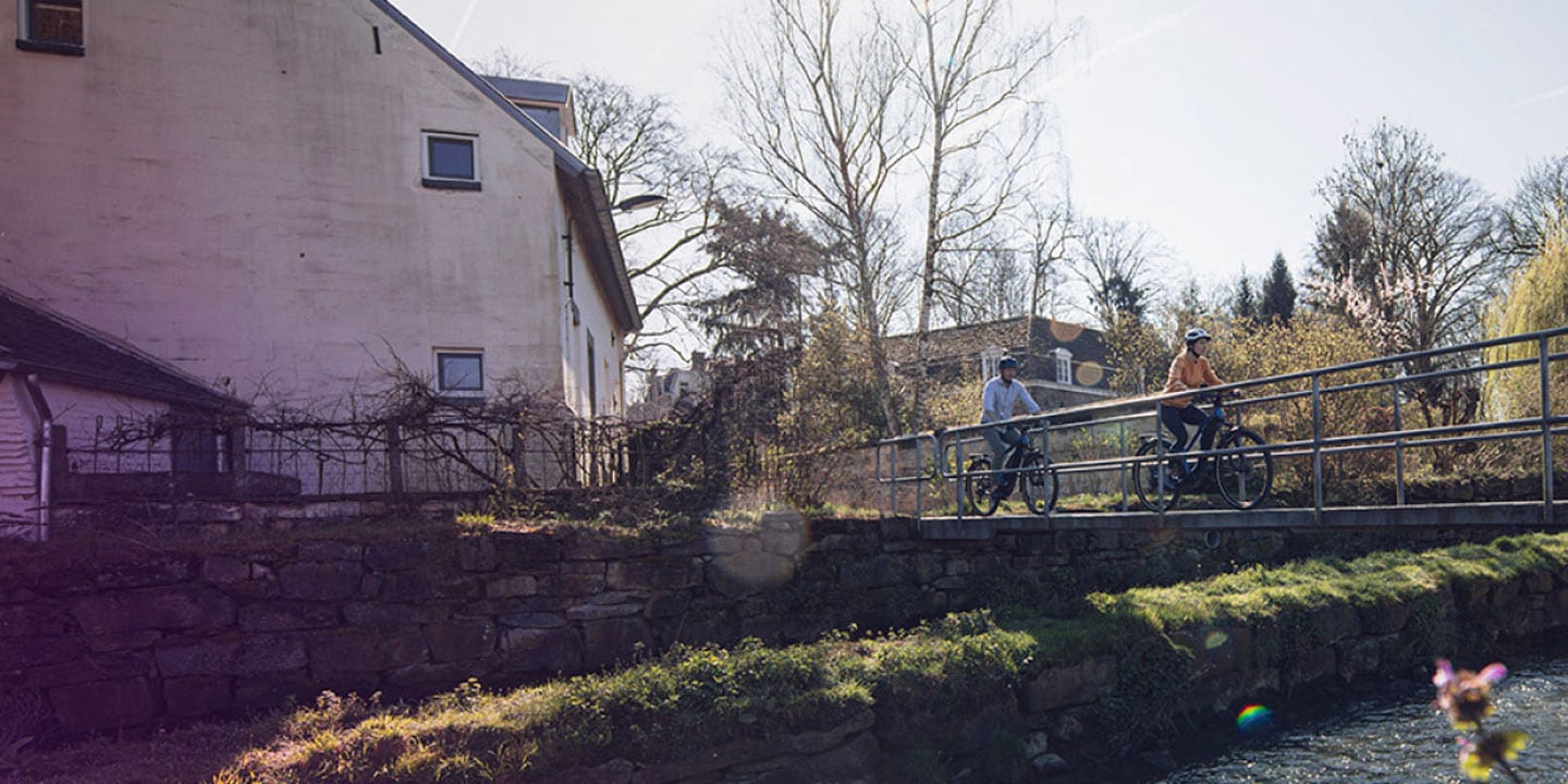 Two cyclists ride across a bridge near a rustic building and a stream on a sunny day