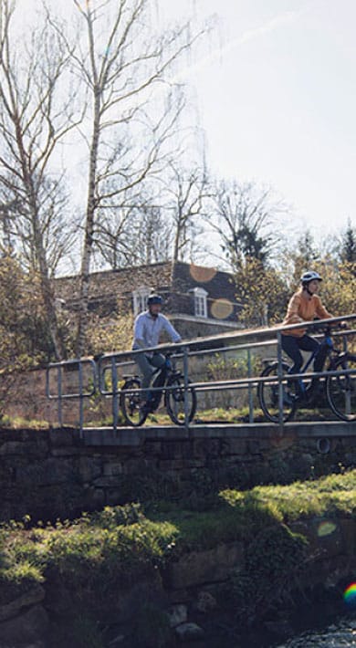 Two cyclists ride across a bridge near a rustic building and a stream on a sunny day