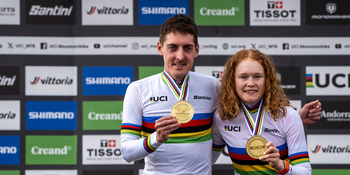 Two cyclists with UCI jerseys pose with gold medals in front of sponsor backdrops.