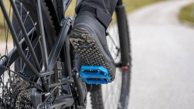 Fuß eines Radfahrers auf einem Mountainbike-Pedal, Schotterweg im Hintergrund.