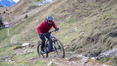 Person fährt Mountainbike auf einem felsigen Pfad mit Hügeln und einem Haus im Hintergrund.