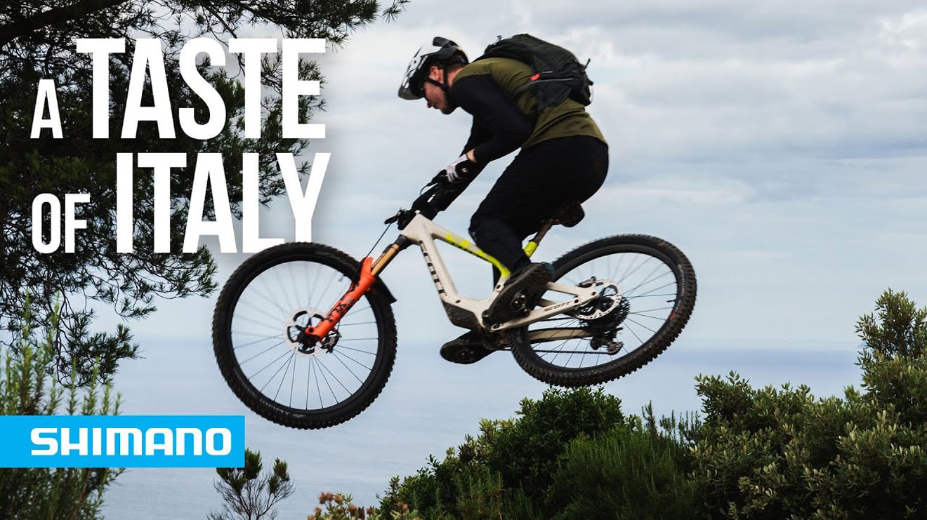 Cyclist mid-air on mountain bike with 'A Taste of Italy' text and Shimano logo.