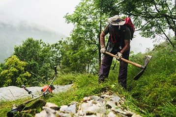 Person clearing grass with a pickaxe on a misty mountain trail.