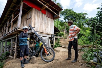 Two men talking outside a wooden house, one with a bike, the other in a striped shirt and hat