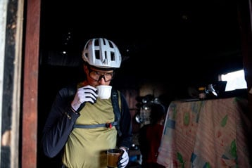 Cyclist wearing a helmet and gloves sipping a drink from a cup inside a dimly lit room.