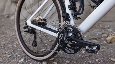 Detailaufnahme des Shimano GRX-Antriebs eines weißen Gravelbikes mit schwarzer Kurbelgarnitur, Kette und Kassette. Das Fahrrad steht auf einem steinigen Pfad.